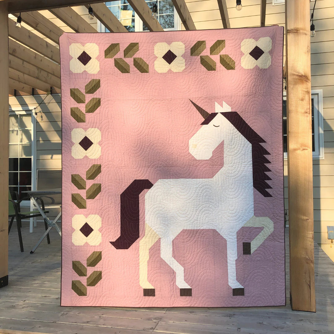 The Unicorn Garden Quilt Pattern by Apples & Beavers features a whimsical pink quilt with a large white unicorn with a brown mane, plus white and green geometric floral borders along the top and left sides, adding extra charm.