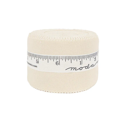 A rolled bundle of Bella Solids Natural Junior Jelly Roll by United Notions, secured with a white ribbon featuring black ruler markings and "moda," is arranged in a cylinder shape against a white background.