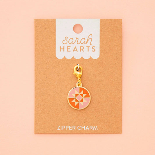 The Riley Blake Round Quilt Star Removable Zipper Charm features a geometric star in pink, peach, and orange with a gold clasp, displayed on a brown card against a peach background.