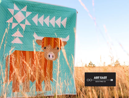 A handmade quilt called "Cattle Call - A Highland Coo Quilt Pattern" with a geometric cow and white triangles is shown in a sunlit grassy field. A black label reads “ART EAST QUILTING CO.”, highlighting this charming farmhouse design.