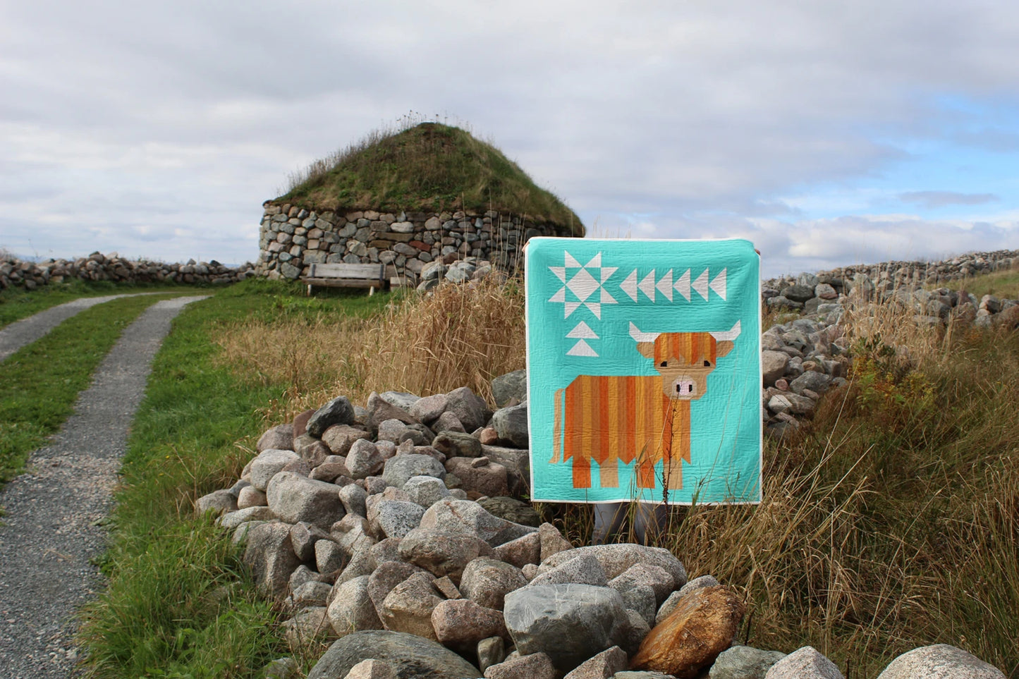 The Cattle Call - A Highland Coo Quilt Pattern features vibrant geometric designs, shown draped over a stone wall by a grassy path, with a stone and grass-roofed building in the background beneath a cloudy sky.