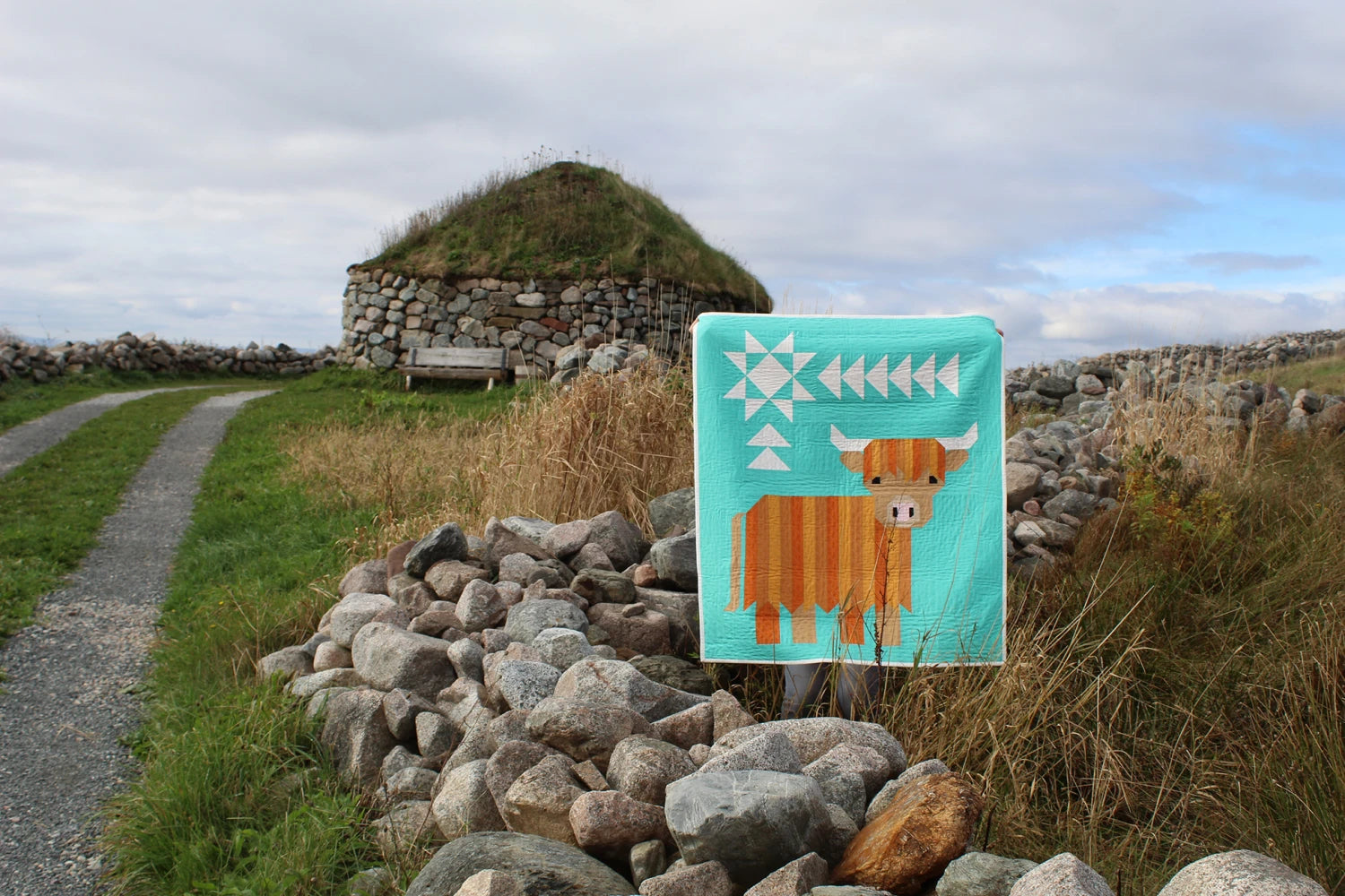 The Cattle Call - A Highland Coo Quilt Pattern features vibrant geometric designs, shown draped over a stone wall by a grassy path, with a stone and grass-roofed building in the background beneath a cloudy sky.