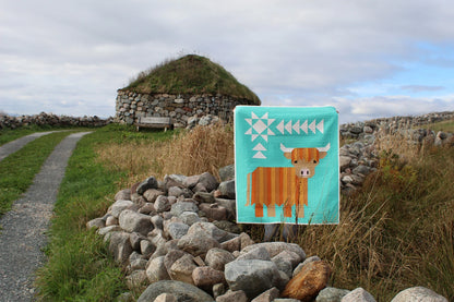The Cattle Call - A Highland Coo Quilt Pattern features vibrant geometric designs, shown draped over a stone wall by a grassy path, with a stone and grass-roofed building in the background beneath a cloudy sky.