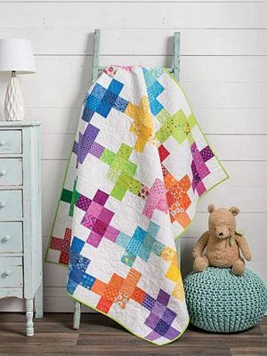 A vibrant cross-pattern quilt from Brewer Sewing's Fast & Fun Quilts for Kids hangs on a wooden ladder beside a white dresser, green pouf, and teddy bear, creating a cozy display against a white shiplap wall.