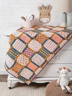 A colorful quilt made with Brewer Sewing’s Fast & Fun Quilts for Kids patterns in pink, blue, mustard, and cream drapes over a white dresser. A giraffe basket with towels and decor sits on top; a plush giraffe toy rests on a beige stool nearby.