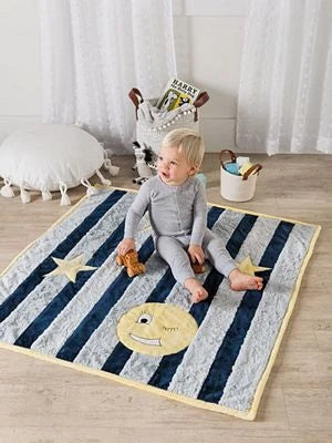 A toddler in gray pajamas sits on a striped blanket with stars and a smiling moon, holding a stuffed animal. Brewer Sewing's Fast & Fun Quilts for Kids adds charm to the cozy room, which also features a round white pillow and basket.