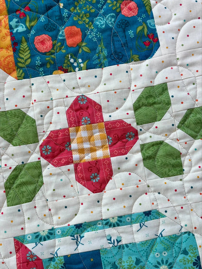 The Novel Idea Quilt Pattern by Riley Blake features geometric floral, gingham, and polka dot fabrics in green leaf shapes and colorful blocks, stitched with swirling lines—perfect for book lovers or pairing with Orchard House fabric.