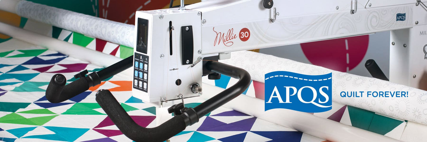 An APQS Demo Millie 30" Long Arm Quilting Machine works on a vibrant geometric quilt, with the APQS logo and “Quilt Forever!” slogan displayed to the right of the machine.
