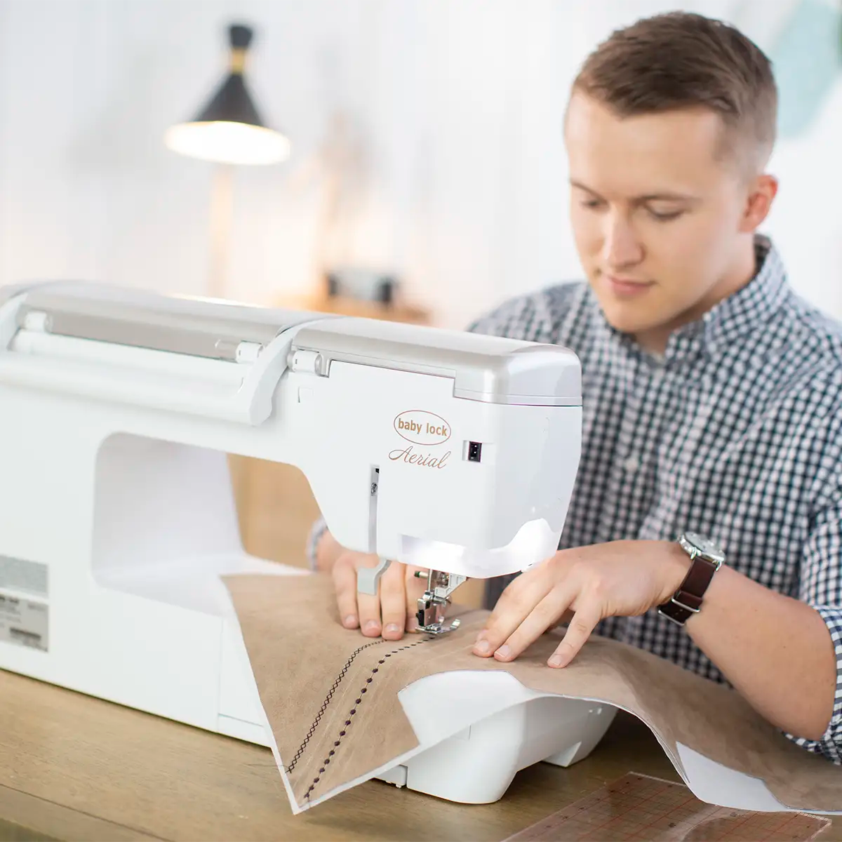 A person in a checkered shirt practices hooping techniques at a table, using the Baby Lock Embroidery 1 Pixie Playdate machine to stitch decorative patterns on brown fabric.