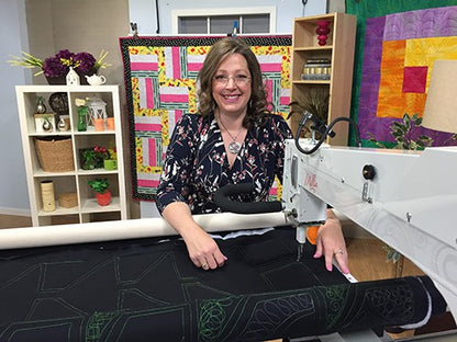 A smiling woman uses the APQS Roadshow by Quilted Joy to quilt a black piece with green stitching. Finished, colorful quilts hang behind her, creating a vibrant display among baskets, plants, and books in her quilting studio.