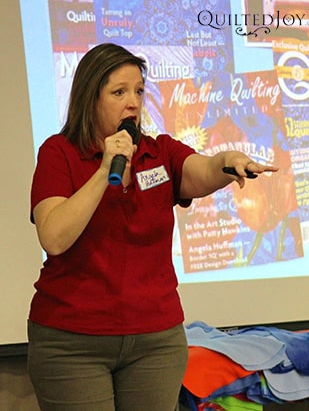 A woman in a red shirt holds a microphone and gestures while speaking in front of a colorful projected presentation. The screen behind her displays quilting-related text and images.