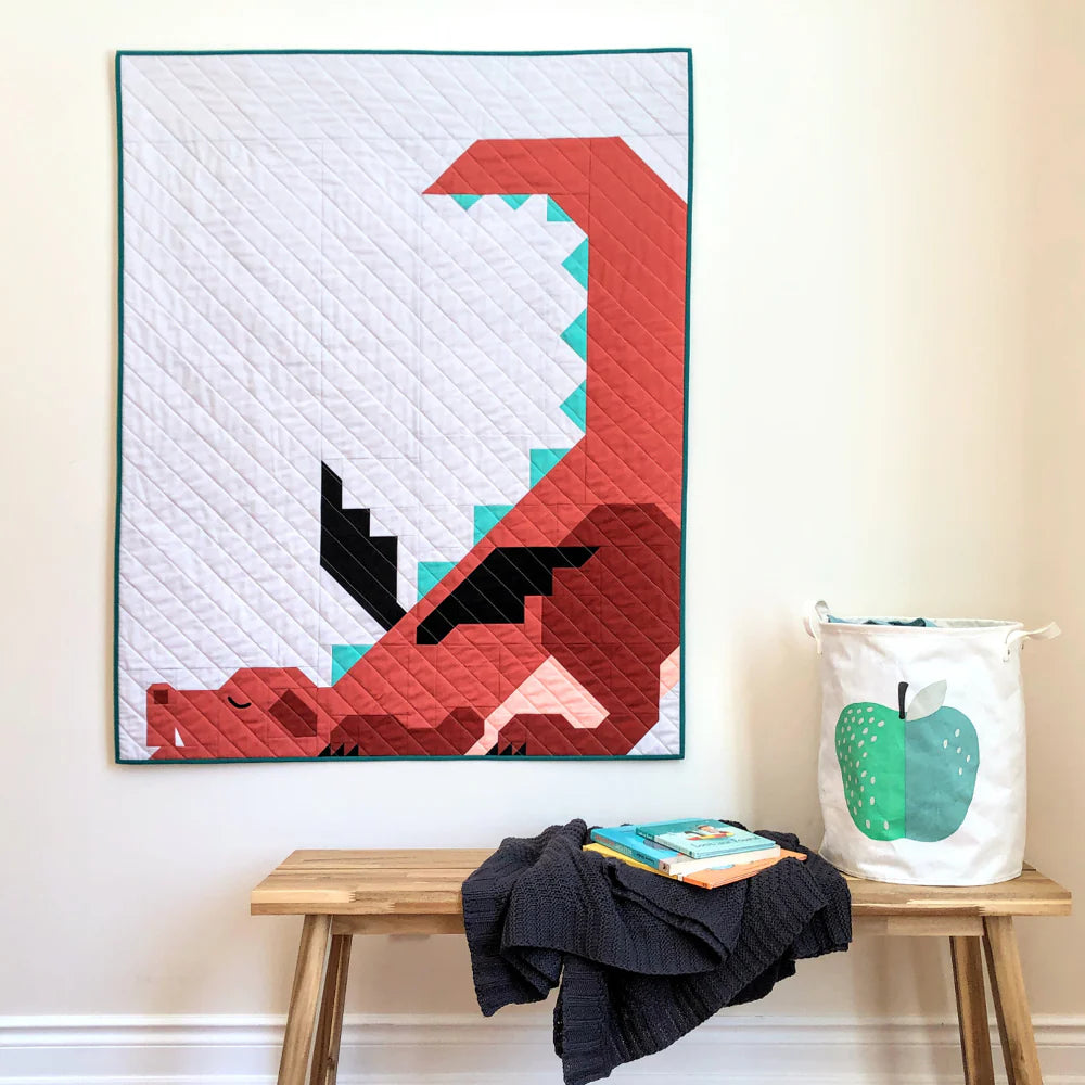 A vibrant quilt made with the Dragon Dreams Quilt Pattern by Apples & Beavers hangs above a wooden bench holding a folded gray blanket and books. Nearby sits a white laundry basket featuring a bold green apple print.