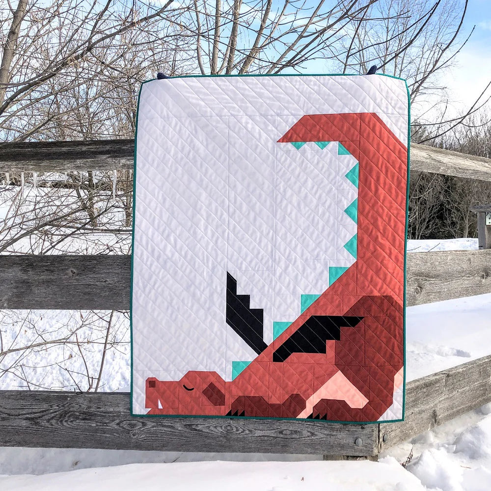 A crib-size quilt featuring a geometric red dragon, made with the Dragon Dreams Quilt Pattern by Apples & Beavers, is draped over a wooden fence on a snowy day, surrounded by leafless trees and snow-covered ground.