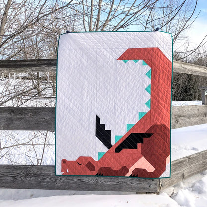 A crib-size quilt featuring a geometric red dragon, made with the Dragon Dreams Quilt Pattern by Apples & Beavers, is draped over a wooden fence on a snowy day, surrounded by leafless trees and snow-covered ground.