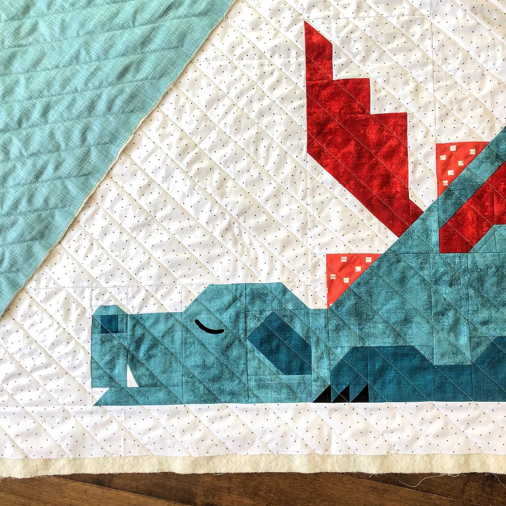 The Dragon Dreams Quilt Pattern by Apples & Beavers features a blue geometric dragon with red spiked wings and closed eyes on a light blue background. Diamond quilting lines add modern style—perfect for crib-size quilts.