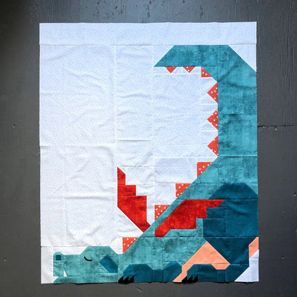 The Dragon Dreams Quilt Pattern by Apples & Beavers features a crib-size quilt block with a geometric dragon of teal, red, and orange on white, showcasing back spikes, a curled tail, and resting eyes.