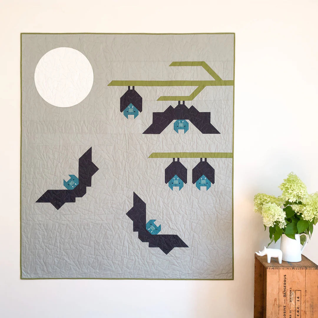 The Bat Buddies Quilt Pattern by Apples & Beavers features geometric bats on a pale backdrop with a full moon and green branch. Ideal for Halloween décor, it’s pictured beside a wooden crate, white vase, and green flowers.