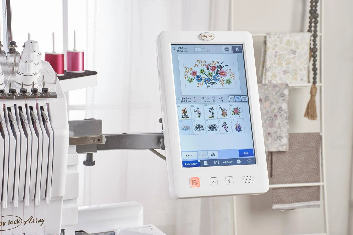 A close-up of the Baby Lock Array 6 Needle Embroidery Machine’s touchscreen shows vibrant floral embroidery designs, with pink thread spools and patterned fabric swatches nearby in a bright, well-lit room.