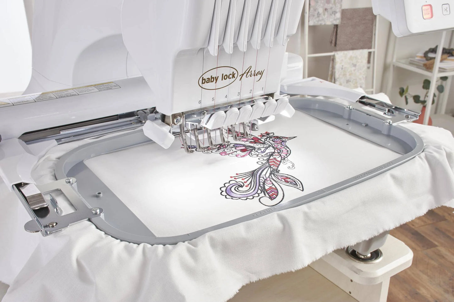 The Baby Lock Array 6 Needle Embroidery Machine is stitching a colorful, intricate floral and bird design onto white fabric in a large hoop, using advanced features for precise embroidery.
