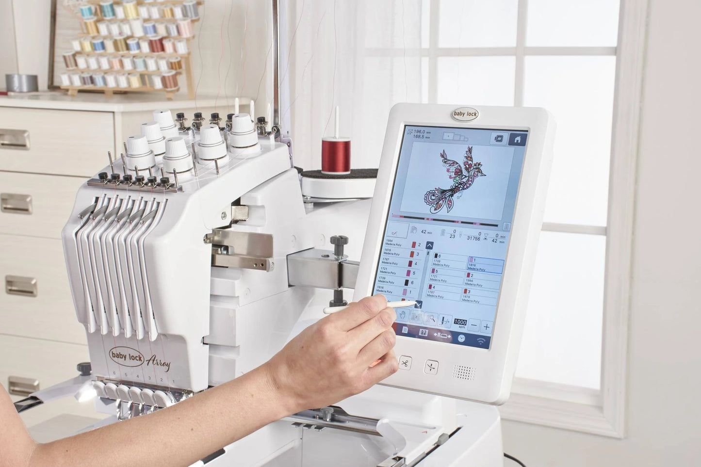 A person operates the Baby Lock Array 6 Needle Embroidery Machine, selecting a vibrant bird design on its touchscreen. Spools of thread and a window are visible in the background of this advanced embroidery device.