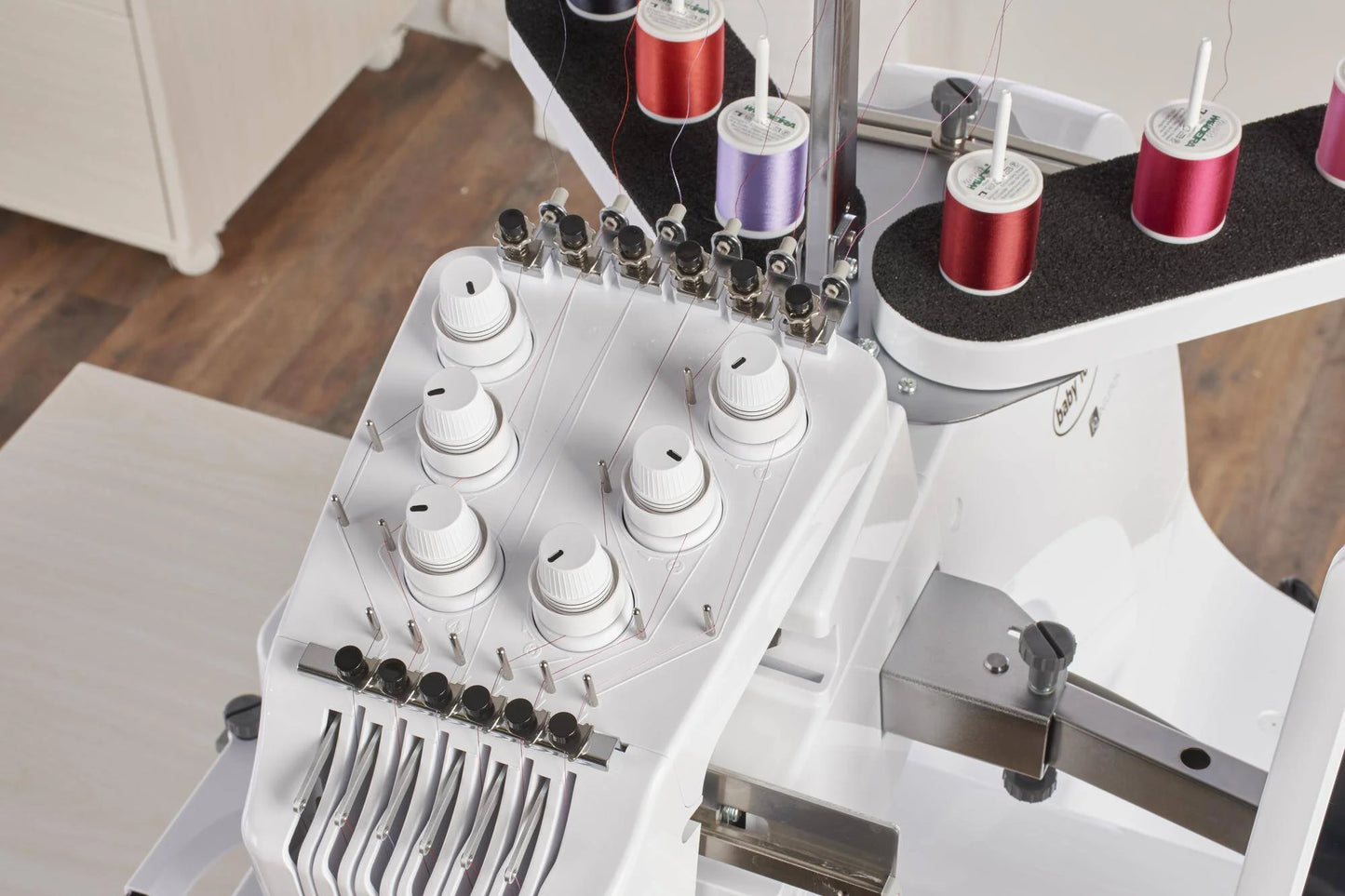 Close-up of the Baby Lock Array 6 Needle Embroidery Machine, showing white dials, silver threading, and colorful spools like purple and red. Perfect for precise embroidery, it’s displayed on a wooden floor.