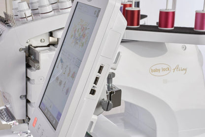 Close-up of the Baby Lock Array 6 Needle Embroidery Machine, featuring its touchscreen display with floral designs, USB ports, and multiple colored thread spools in the background.