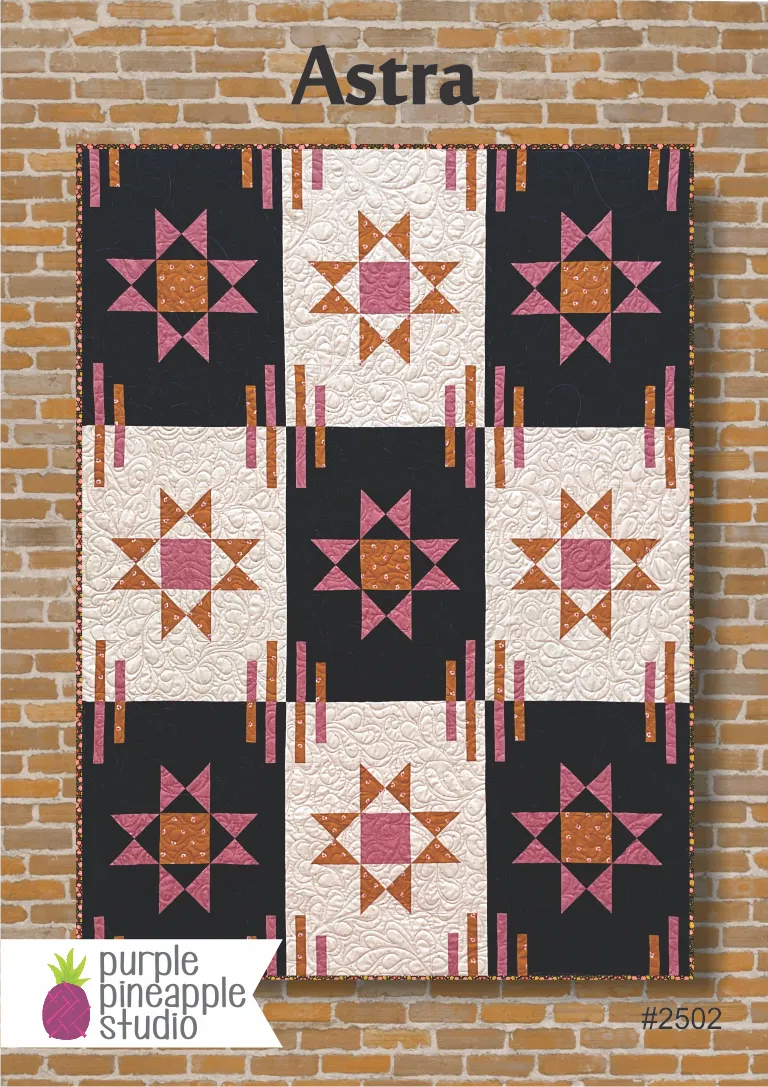 The Astra Quilt Pattern by Purple Pineapple Studio hangs against a brick wall, displaying star motifs in pink, orange, and white on black and white squares. The Purple Pineapple Studio logo appears in the bottom left corner.