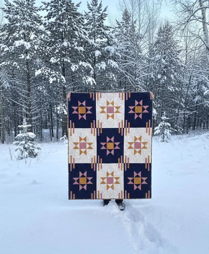 A person stands in a snowy forest holding up the Purple Pineapple Studio Astra Quilt Pattern, which features star designs in navy blue, gold, and cream, with only their hands and feet showing.