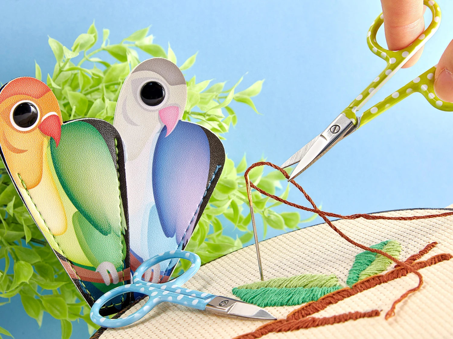 A hand threads a needle over an embroidery hoop using Baby Lock Purse Parrot Embroidery Scissors With Pouch, featuring green polka dots and stainless steel blades. Nearby, blue scissors rest in a colorful bird-shaped holder against a bright background.