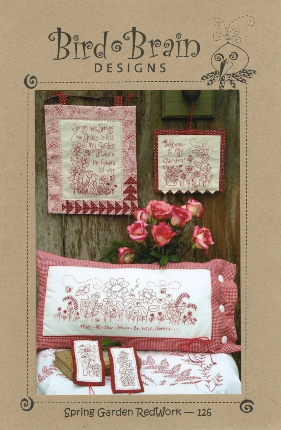A display of Checker Distributor’s Spring Garden Redwork Quilt Pattern features floral embroidery for wall hangings, a pillow, and bookmarks with garden-themed phrases, all set beside a vase of pink roses.
