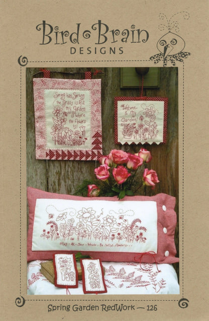 A display of Checker Distributor’s Spring Garden Redwork Quilt Pattern features floral embroidery for wall hangings, a pillow, and bookmarks with garden-themed phrases, all set beside a vase of pink roses.