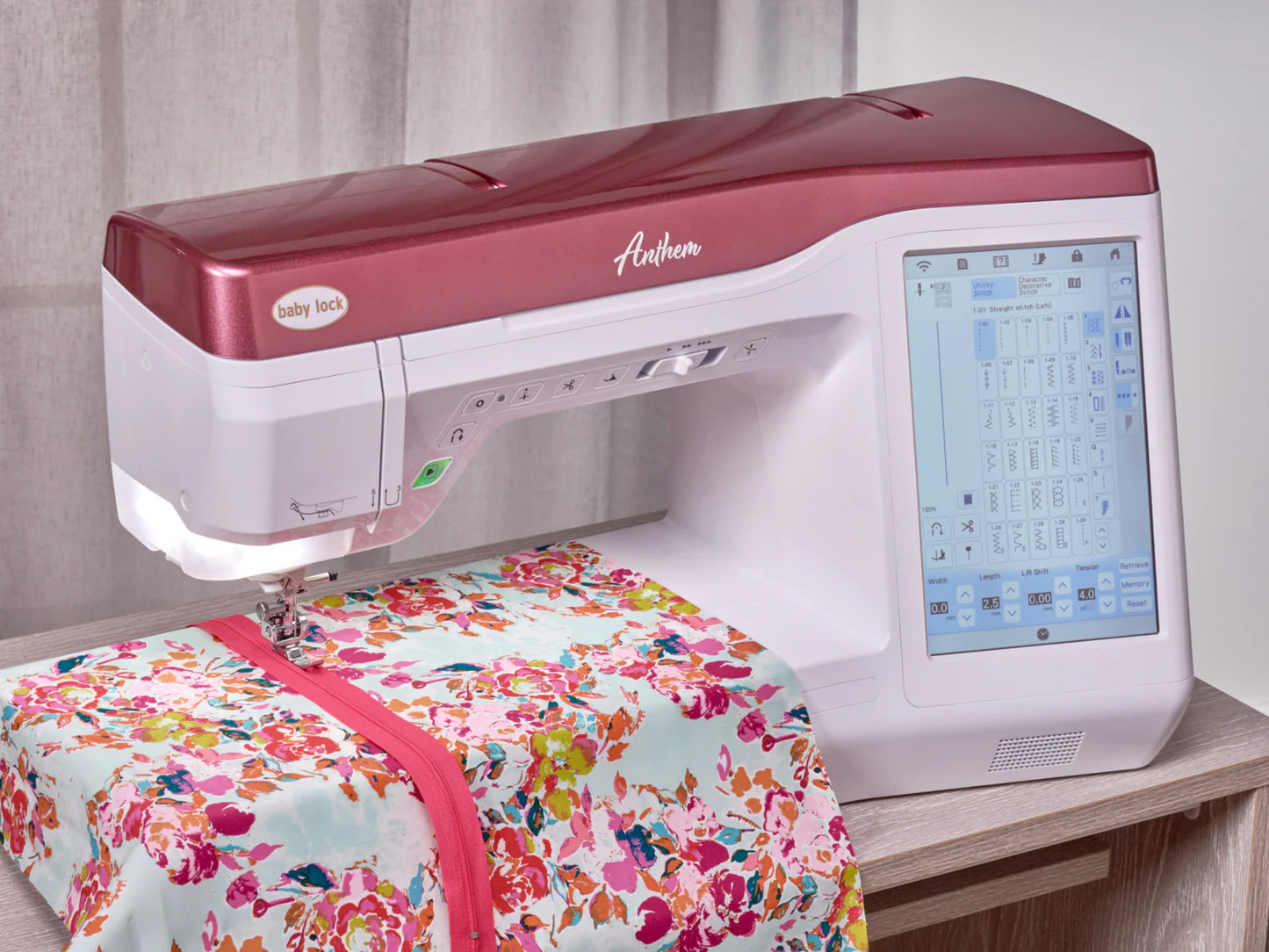 A Baby Lock Anthem Sewing & Quilting Machine with FREE 60 Days of Online Classes is stitching colorful floral fabric trimmed with pink ribbon on a wooden table near curtains.