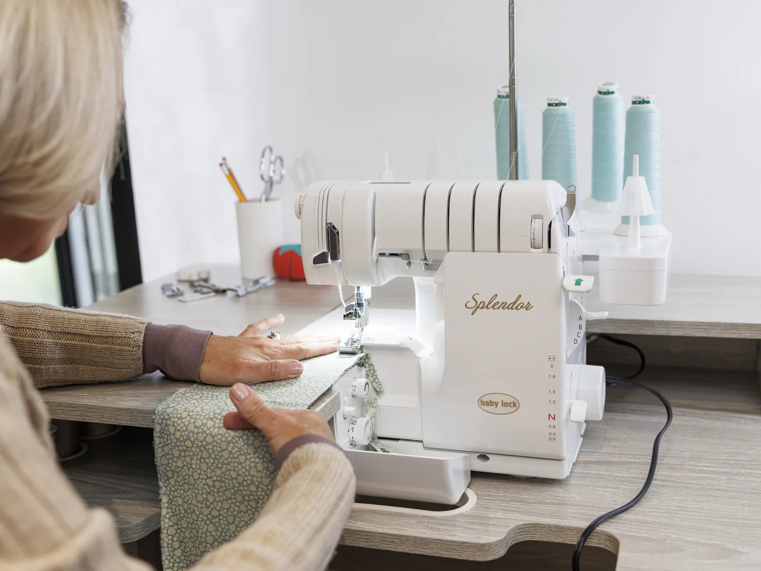 Using the Baby Lock Splendor 8-Thread Combination Serger with ExtraordinAir™ Threading and FREE 60 days of online classes, a person stitches light blue patterned fabric at a wooden table, surrounded by thread spools and sewing tools.