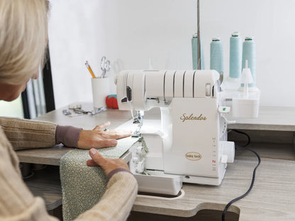Using the Baby Lock Splendor 8-Thread Combination Serger with ExtraordinAir™ Threading and FREE 60 days of online classes, a person stitches light blue patterned fabric at a wooden table, surrounded by thread spools and sewing tools.