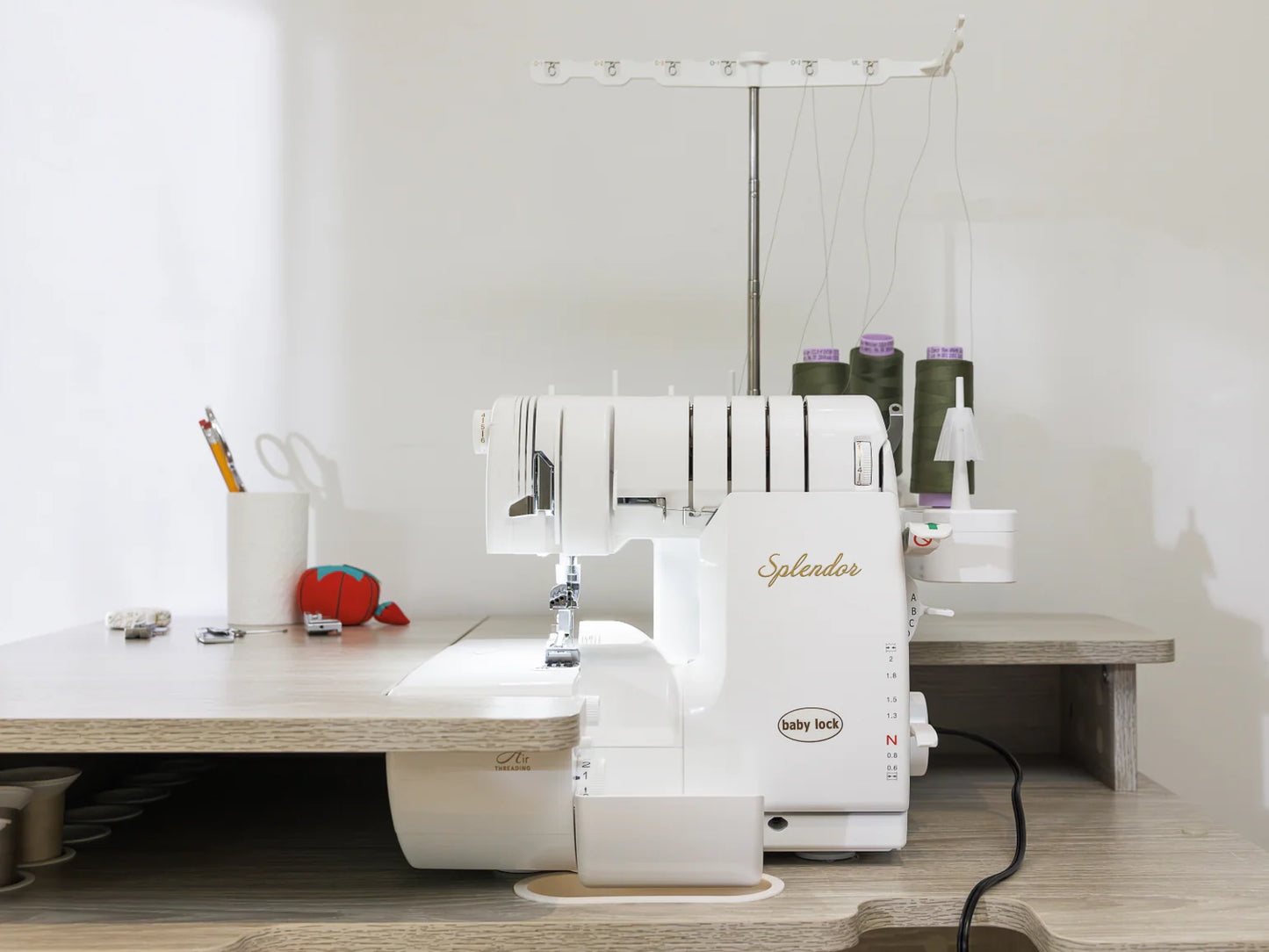 A Baby Lock Splendor 8-Thread Combination Serger with ExtraordinAir™ Threading and FREE 60 Days of Online Classes sits on a wooden table, surrounded by scissors, spools of thread, and a red pincushion.
