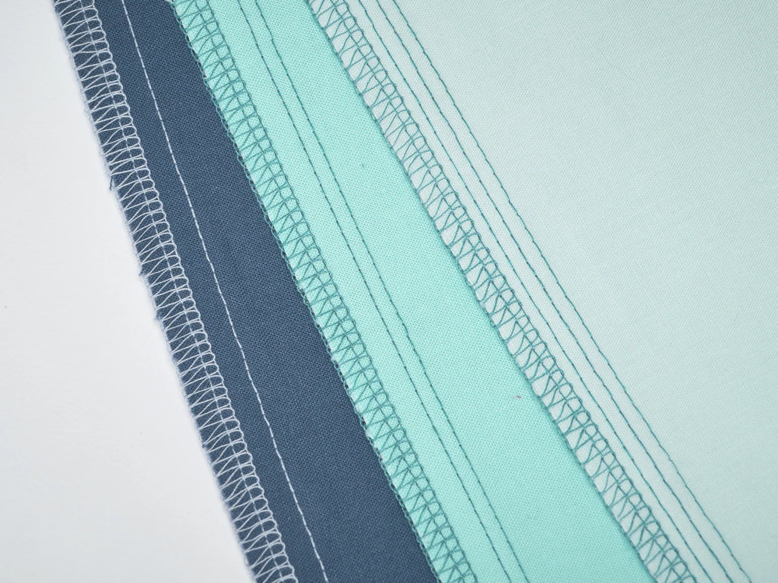 Four blue and aqua fabric pieces are stacked diagonally, showing their stitched edges finished with the Baby Lock Splendor 8-Thread Combination Serger with ExtraordinAir™ Threading on a white background. Includes 60 days of free online classes.