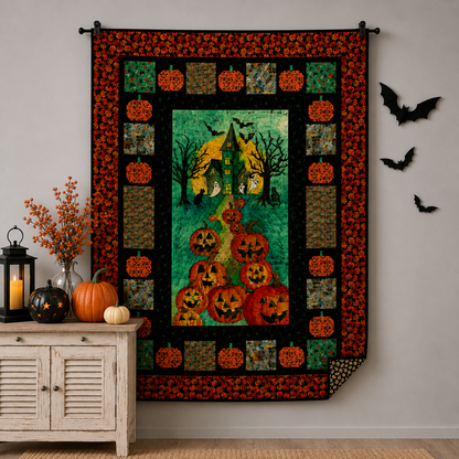 The Bewitched House Quilt Kit by Quilted Joy, displaying pumpkins, jack-o’-lanterns, a haunted house, and bats, hangs on a wall. Next to it are black bat decorations and a table with pumpkins, a lantern, and orange flowers.