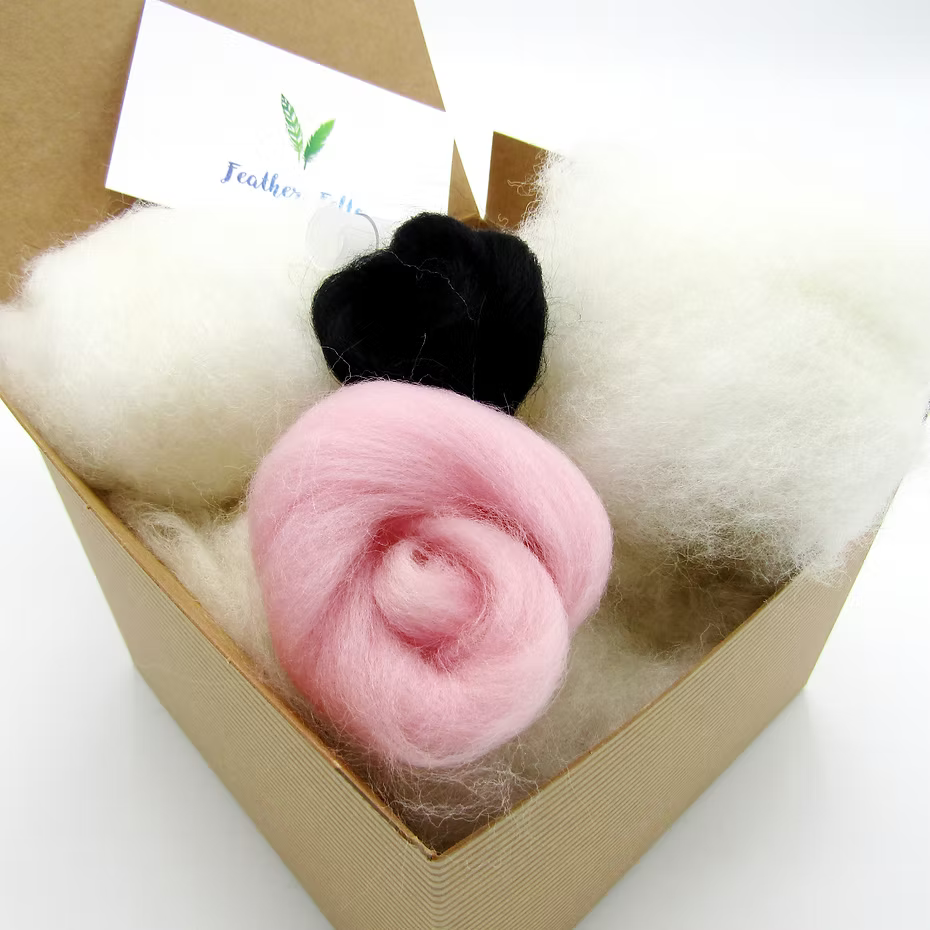 A brown box holds fluffy white, pink (spiral rolled), and black wool for a bunny project from the Feather Felts Bunny - Needle Felting Kit No Foam; a white card with green leaves and blue text is partly visible in the background.