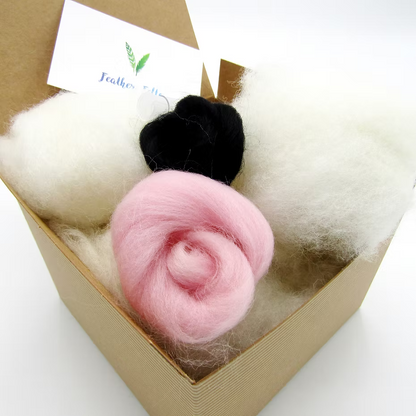 A brown box holds fluffy white, pink (spiral rolled), and black wool for a bunny project from the Feather Felts Bunny - Needle Felting Kit No Foam; a white card with green leaves and blue text is partly visible in the background.