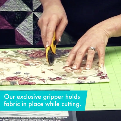 A person uses the Creative Grids Quilt Ruler Circles Set of 5 by Checker Distributor with a rotary cutter to cut floral fabric on a green mat. Text reads, Our exclusive gripper holds fabric in place while cutting. A quilt is seen in the background.