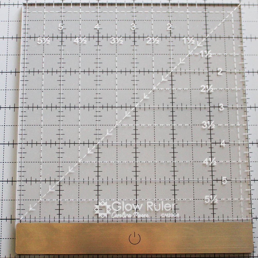 The United Notions Carolina Moore 6" Glow Ruler is a clear illuminated acrylic ruler with white grid lines, measurements, a metal edge, and a power button symbol, shown resting on a gridded mat.