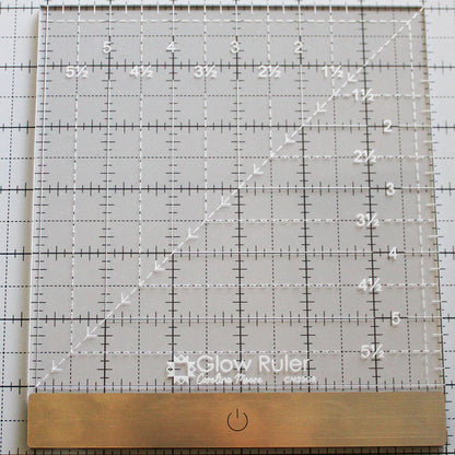 The United Notions Carolina Moore 6" Glow Ruler is a clear illuminated acrylic ruler with white grid lines, measurements, a metal edge, and a power button symbol, shown resting on a gridded mat.