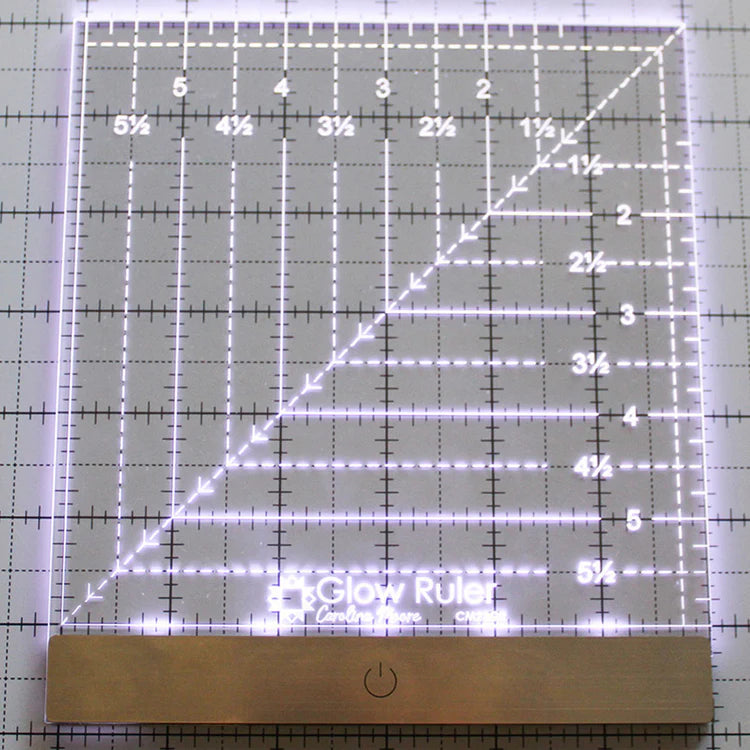 The Carolina Moore 6" Glow Ruler by United Notions is an illuminated acrylic quilting ruler with grid lines, measurements from 1 to 6.5 inches, edge lighting, and a sleek metal power button at the base.