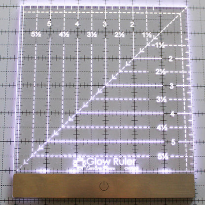 The Carolina Moore 6" Glow Ruler by United Notions is an illuminated acrylic quilting ruler with grid lines, measurements from 1 to 6.5 inches, edge lighting, and a sleek metal power button at the base.