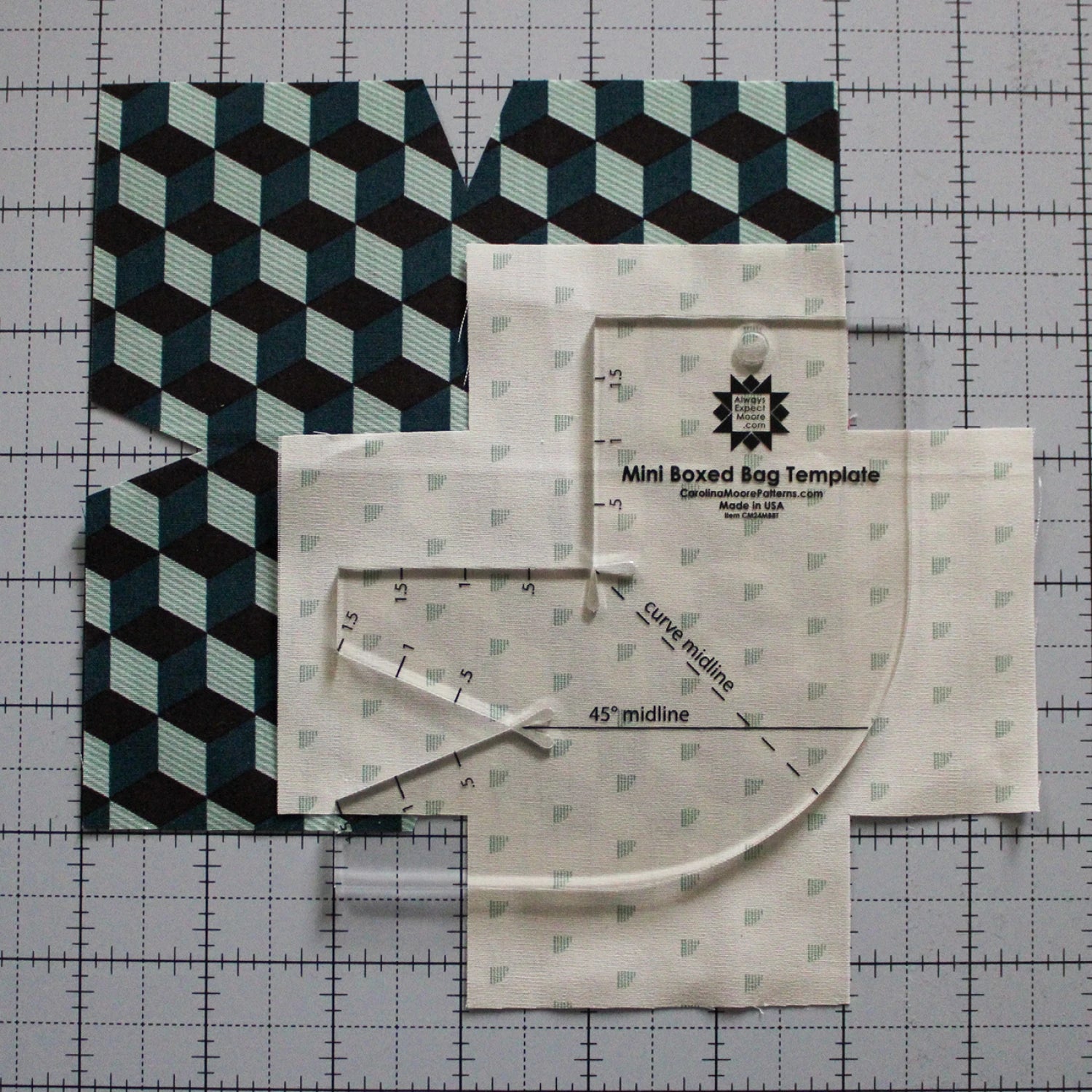 Four geometric cube and three small diamond fabric pieces are arranged on a cutting mat with the Checker Distributor Mini Boxed Bag Template, a clear acrylic tool for precise bag sewing pattern projects.