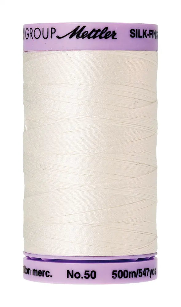 A spool of Mettler Cotton Silk Finish 50wt 500m Candlewick quilting thread in light cream, featuring a purple and white label and containing 500 meters of cotton sewing thread.
