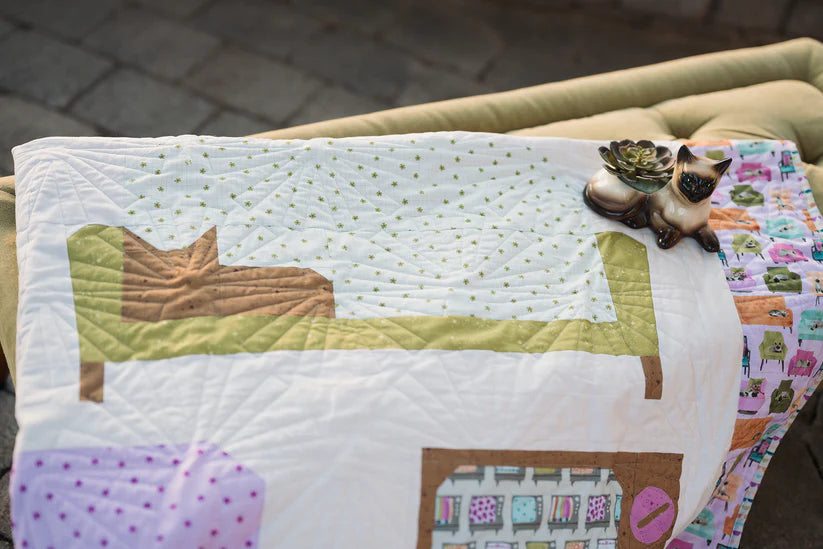 The Couch Potatoes Quilt Pattern by Checker Distributor, featuring pastel geometric designs and a cat on a bed, is draped over a green couch with a small cat-shaped ceramic planter on top.