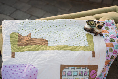 The Couch Potatoes Quilt Pattern by Checker Distributor, featuring pastel geometric designs and a cat on a bed, is draped over a green couch with a small cat-shaped ceramic planter on top.