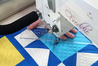 A user quilts a geometric pattern on a blue, yellow, and white quilt with the APQS Demo Freddie 26" Long Arm Quilting Machine by APQS, guiding the fabric by hand with a transparent ruler.