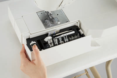 A hand opening the accessory storage built into the auxiliary bed of the JUKI Hayate DX-4500QVP, with presser feet inside.